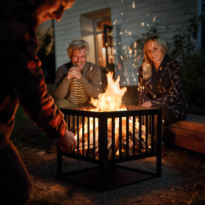Three people gathered around a square black steel slatted fire pit burning logs in a garden at night outdoor wood burning fire pit Pyrova