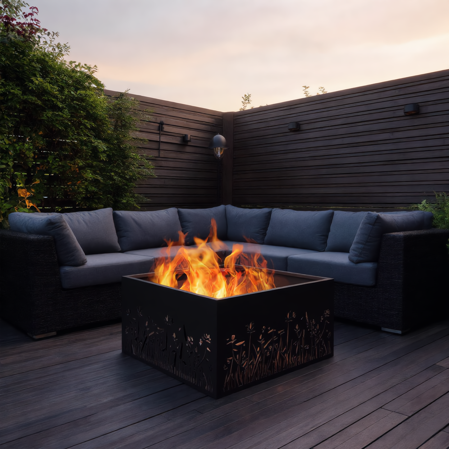 Black steel square fire pit with floral cut-out panels burning wood at dusk on decking terrace next to grey rattan corner sofa outdoor fire pit Pyrova